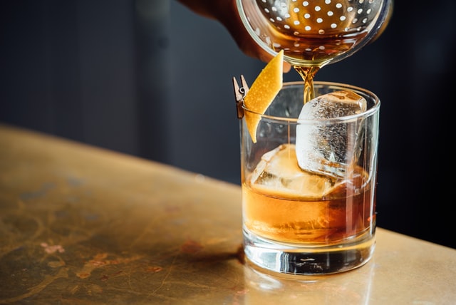 Whiskeyglas med två stora isbitar i, på kanten ett apelsinskal, Från shakern ovanför hälls det i en brun orange dryck