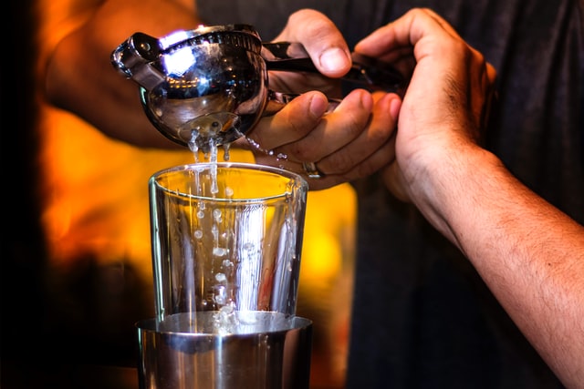 Två händer som pressar ihop en rostfri citruspress. Citruspressen utger vätska som droppar ner i ett transparent glas