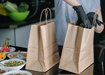 Diskbänk med två bruna papperspåsar där person med svart handskar lägger i mat förpackningar för take away