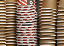 Travar med pappersmuggar för engångsbruk. En variant med röd och vit design, placerade bredvid bruna kaffemuggar. 