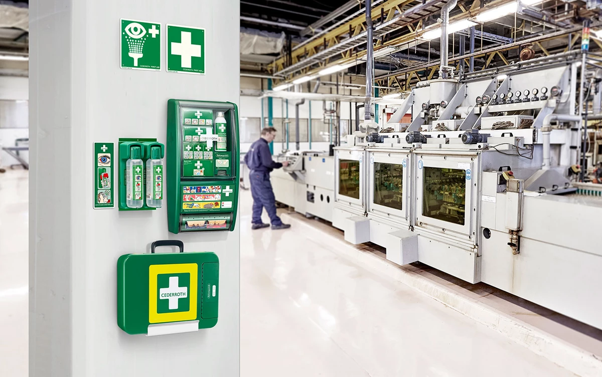 Första hjälpen utrustning monterad på pelare i en industrihall med maskiner och en arbetare i bakgrunden.