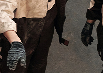 Två personer sett från midjan och ner, iklädda mörka kläder och grå handskar, stående på ett betonggolv. 