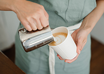  Person som håller en rostfri behållare med skummad mjölk som hälls i en vit dryckesbehållare i papper som innehåller kaffe.