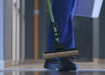 Person i blåa byxor och svarta skor kommer gåendes i en korridor hållandes i en mopp med grönt handtag