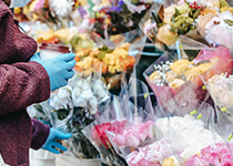 En person i en vinröd kappa och blå handskar väljer ut blommor inslagna i cellofan från en färgglad blomstermarknad utomhus.