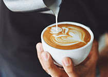 En person på ett café häller skummad mjölk i en kopp kaffe, vilket skapar en hjärtform i kaffekoppen.