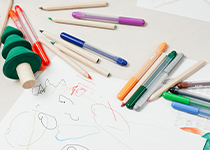 Färgglada skolmaterial och pennor utspridda på en teckning med enkla former och linjer, på en vit yta med staplingsleksaker.
