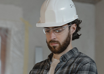 En fokuserad byggnadsarbetare med vit hjälm, skyddsglasögon och rutig skjorta tittar noga på något utanför kameran i en arbetsmiljö.