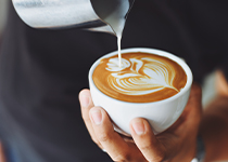 Rostfri tillbringare fylld med skummad mjölk som hälls ner i en vit kopp fylld med kaffe och skapar mönster. Koppen hålls utav en hand