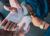 En hand som blir omlindad med ett vitt bandage av en annan person. Den som blir omlindad har en blå filt runt sig och en klocka på andra handen.