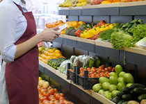 Person i sina arbetskläder, blå skjorta och rött förkläde i profil mot en hylla med frukt och grönsaker prydligt upplagt bredvid varandra på flera hyllplan