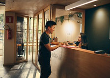 En man interagerar med en receptionist vid en modern reception på ett hotell. Interiören har varm belysning och inredning i trä.