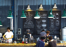 Cafe Miljö med högt i tak och stora gröna lampor som hänger ner över disken, i bakgrunden ser man stora svarta griffeltavlor med meny