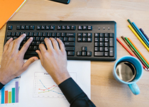 En persons händer skriver på ett svart tangentbord vid ett skrivbord i trä. Det finns färgpennor, ett diagram och en kopp kaffe på bordet.