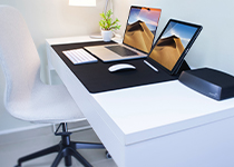 Ett modernt, rent kontorsbord med en surfplatta, en bärbar dator, tangentbord, mus, extern hårddisk och en liten krukväxt placerad mot en blå vägg.