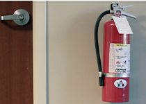 En röd brandskydd monterad på en beige vägg nära en stängd dörr med ett metalliskt dörrhandtag. 