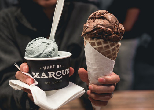 En person står vid en kiosk och håller i två glassar, en i en strut och den andra i en kopp märkt "Made by Marcus". Bakgrunden är i oskärpa. 